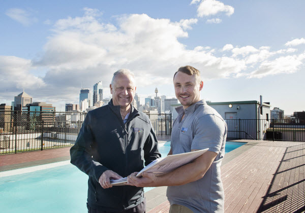 Two men standing in front of a pool on a roof and smiling at the camera while holding a book